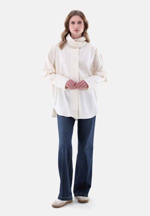 Camicia oversize color crema con bottoni e maniche lunghe, abbinata a jeans svasati blu scuro e sneaker bianche. Design semplice e classico.