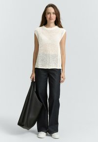 Cremfarbener, gestrickter Kurzarm-Top mit lockerer Passform, kombiniert mit weitem, dunkelblauen Denim-Hosen und weißen Sneakers. Das Model hält eine schwarze Tragetasche.