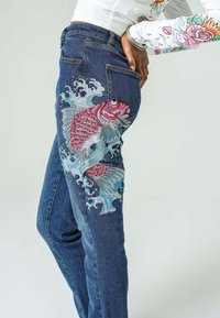 Jeans denim dengan desain ikan koi dan ombak yang dijahit cerah di kaki kiri. Jeans berwarna biru klasik dengan potongan slim.