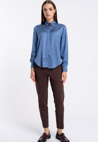 Kobieta ubrana w niebieską satynową bluzkę z długim rękawem i broszką, brązowe wąskie spodnie oraz ciemne bordowe mokasyny, stojąca na tle białym.