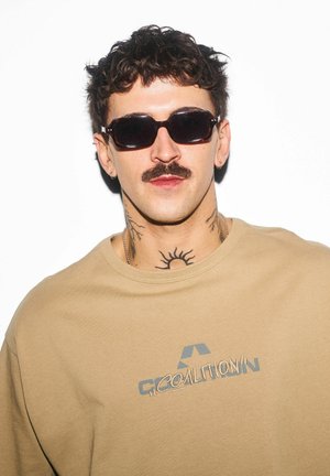 Mężczyzna o ciemnych, kręconych włosach, z wąsami i tatuażem słońca na szyi, noszący ciemne okulary przeciwsłoneczne i beżową bluzę "Coalition".