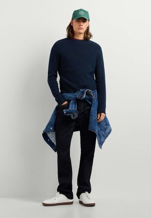 Junger Mann mit grüner Mütze, marineblauem Pullover, dunklen Jeans, weißen Sneakers und einer Jeansjacke, die um die Taille gebunden ist, steht vor einem einfarbigen Hintergrund.