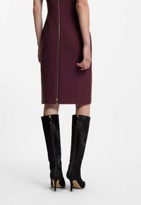 Burgundin väriin puettu kapea mekko, jossa on näkyvä vetoketju selkäpuolella, yhdistettynä polven ylle ulottuviin mustiin mokkasaappaisiin, joissa on sivuvetoketjut ja korkeat korot.