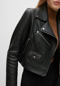 Sort læder cropped bikerjakke med tekstureret overflade, sølv lynlåse, trykknapper og en brystlomme, stylet over en brun top.