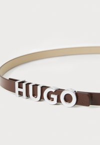 Brauner Leder Gürtel mit silbernen Buchstaben, die "HUGO" buchstabieren. Der Gürtel hat eine glatte Textur und ein minimalistisches Design.