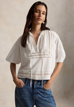 Femme aux longs cheveux foncés portant un chemisier blanc avec des inserts en dentelle et des manches bouffantes, ainsi qu'un jean bleu avec les mains dans les poches.