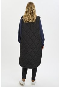 Gilet long matelassé noir avec un col haut, sans manches et un ourlet arrondi, porté par-dessus un haut navy et un pantalon noir. Tissu léger et texturé.