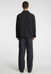 Must jakk kaelusega, nööbitavate varrukaotsade ja sirge lõikega, koos tumedas mereväes siniste pükstega, vaadatuna tagantpoolt.