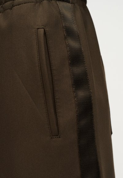 Τα καφέ παντελόνια διαθέτουν πλαϊνές τσέπες και μια αντίθετη μαύρη σατέν ρίγα κατά μήκος της πλευράς, αναδεικνύοντας μια λεία υφή υφάσματος.