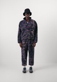 Purple and black patterned fleece jacket with a high collar and half zip, paired with matching pants. Accessories include a black bucket hat and sunglasses.