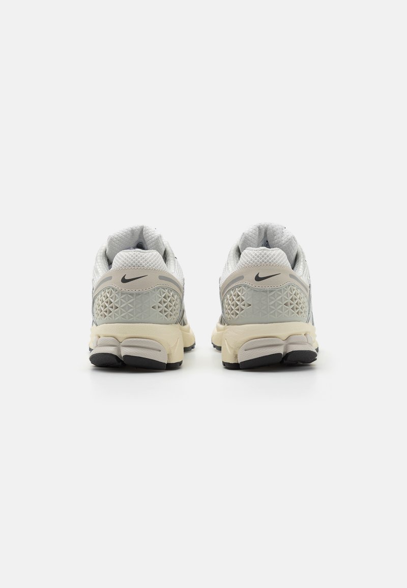 Para białych i beżowych sneakersów Nike pokazana z tyłu, z czarnym logo, teksturowanym obcasem oraz grubymi czarnymi i beżowymi podeszwami.