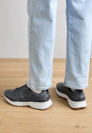 Jeans azzurri chiari e sneakers nere con suole bianche indossate su pavimento in legno, viste da dietro all'altezza della caviglia.