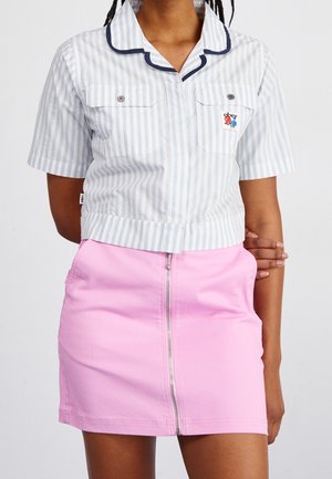 Donna che indossa una camicia a maniche corte a righe azzurro chiaro e bianco con colletto e tasche sul petto, abbinata a una minigonna rosa con zip frontale.
