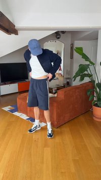Person som har på seg en blå caps, marineblå shorts, hvite joggesko og en mørk jakke, står i en stue med en brun sofa og en stor grønn plante.