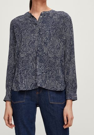 Femme portant un chemisier bleu marine à manches longues avec un motif abstrait blanc et un jean bleu foncé, se tenant devant un fond uni.