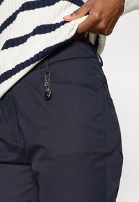 Sötétkék nadrág vékony fitneszes szabású, zipzáros zsebbel és egy kis fémcharmmal a zipzáron. Az anyag sima és tartós hatású.