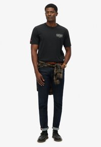 Camiseta de algodón negra con el logo de "Superdry" en el lado izquierdo del pecho, combinada con jeans de mezclilla oscura y una camisa de franela a cuadros atada alrededor de la cintura.