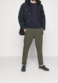 Marinblå puffjacka med pälsfodrad huva, mörk tröja, olivgröna byxor och blå skor med ljusa sulor. Minimala accessoarer synliga.