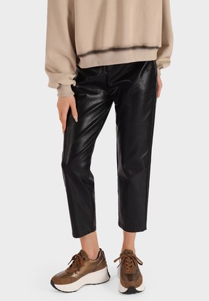Pantaloni in ecopelle nera con una texture lucida, di lunghezza cropped, abbinati a sneaker marroni chunky. Un morbido felpa beige completa l'outfit.