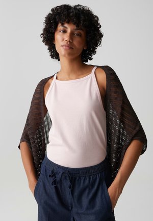Giovane con capelli corti e ricci, indossa un top rosa chiaro senza maniche, uno scialle scuro a maglia aperta e pantaloni blu navy con coulisse, mani nelle tasche.