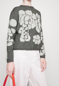 Personne portant un pull gris avec de grands motifs floraux blancs et un pantalon rose clair, tenant un sac à main à bandoulière rouge sur le côté.