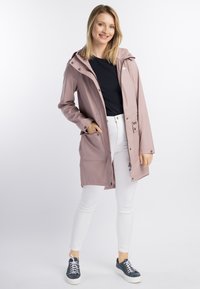 Leende kvinna iklädd en ljus mauve regnjacka över svart topp, vita byxor och marinblå sneakers, stående med handen i fickan mot en vit bakgrund.