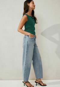 Mujer con una blusa verde sin mangas, jeans de cintura alta de color azul claro con cuentas en los bordes de los bolsillos, y sandalias de tacón negras, se encuentra de pie contra una pared sencilla.