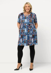 Sininen kuviollinen mekko lyhyillä hihoilla, jossa on geometrisiä ja heimoteemaiset kuviot, yhdistetty mustiin legginsseihin ja korkokenkiin.
