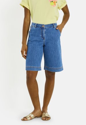 Personne portant un short en denim bleu mi-long, un t-shirt jaune pâle avec un motif estival, et des sandales dorées à enfiler, debout avec une main dans la poche.