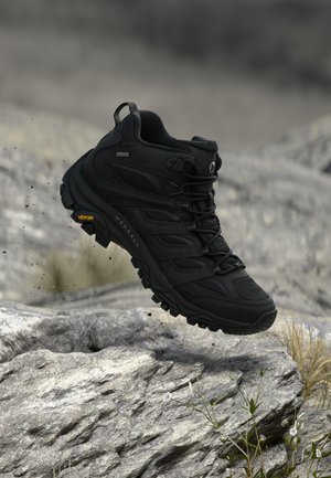 Μαύρο πεζοπορικό παπούτσι με σόλα Vibram που αιωρείται πάνω από βραχώδες έδαφος με μικρά φυτά και ξερό χορτάρι στο φόντο.
