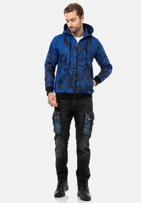 Kapuze mit Reißverschluss in Blau mit schwarzem grafischem Muster, kombiniert mit schwarzen Cargo-Hosen mit blauen Denim-Akzenten und mehreren Taschen.