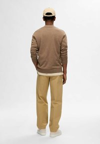 Brauner Strickpullover mit gerippten Bündchen, kombiniert mit beigen lockeren Hosen und hellen Schuhen, von hinten gegen einen schlichten Hintergrund gesehen.