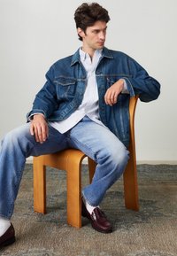Veste en denim avec des boutons, chemise rayée bleu clair, jeans bleu clair et mocassins bordeaux. Chaise en bois sur un sol texturé.