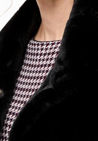 Sort faux-fur krave af et beklædningsstykke med et nærbillede af halsområdet, der har et burgunder og hvidt hundetand mønster nedenunder.