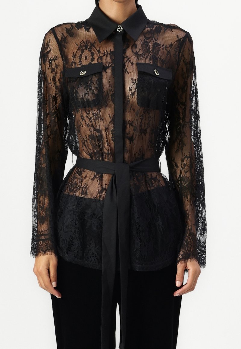 Blouse noire en dentelle à manches longues avec motif floral, col uni, deux poches poitrine avec boutons et ceinture nouée à la taille sur pantalon noir.