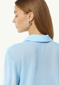 Jasnoniebieska, półprzezroczysta bluza z kołnierzykiem, charakteryzująca się gładką fakturą i zwiewnym krojem. Modelka nosi srebrne kolczyki w kształcie kół.