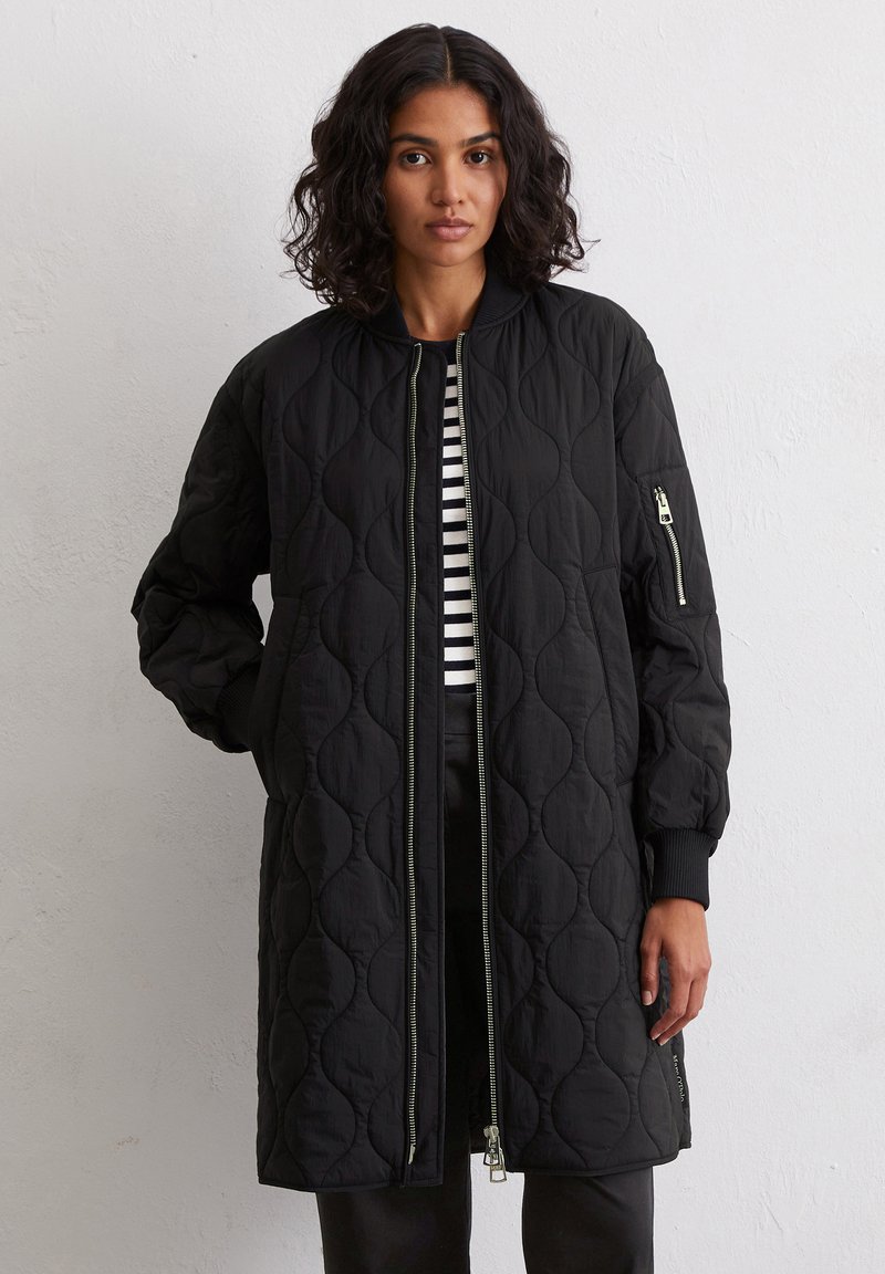 Femme aux cheveux bouclés portant un long manteau noir matelassé par-dessus une chemise à rayures noir et blanc, se tenant devant un mur blanc uni.