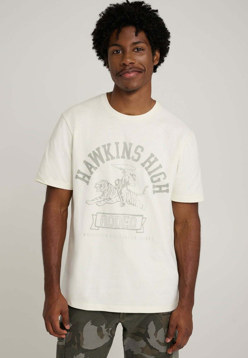Kremowy t-shirt z nadrukiem przedstawiającym jeźdźca rodeo i tygrysa. Krótkie rękawy, okrągły dekolt i miękki bawełniany materiał.