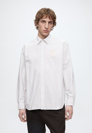 Joven con el pelo ondulado que lleva una camisa blanca de rayas finas con botones y un pequeño parche bordado en el pecho, de pie frente a un fondo liso.