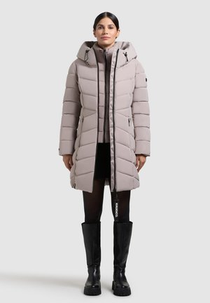 Hellgraue gepolsterte Jacke mit Kapuze, vertikalen Nähten, Reißverschlusstaschen und einem Markenanhänger. Das Modell trägt knielange schwarze Stiefel.