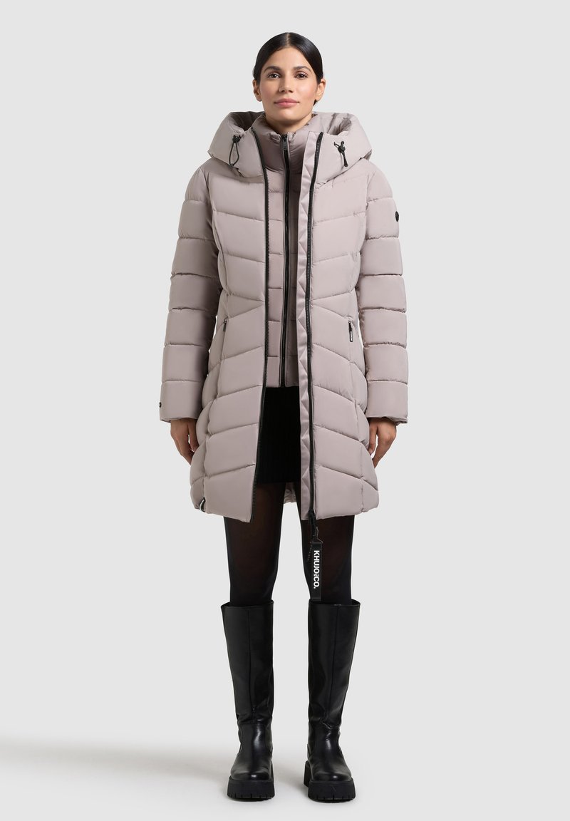 Svetlo sivý prešívaný kabát s kapucňou, s vertikálnym švom, zipsovými vreckami a značkovým ťahátkom. Model má na sebe čierne čižmy po kolená.