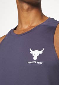 Detailní záběr na osobu v bezrukávovém fialovém tričku s bílým logem býčí lebky a textem "PROJECT ROCK" na hrudi.