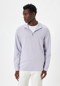 Sudadera con cremallera en lavanda clara, confeccionada en una tela suave, con mangas largas y un cuello sutil. Combinada con pantalones de tonos claros.