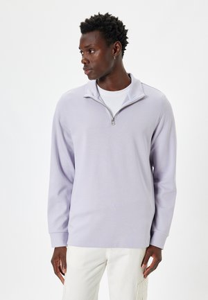 Sudadera con cremallera en lavanda clara, confeccionada en una tela suave, con mangas largas y un cuello sutil. Combinada con pantalones de tonos claros.