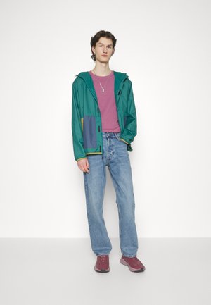 Green and blue windbreaker with a hood, gray accents, over a purple t-shirt, paired with light denim jeans and maroon sneakers.