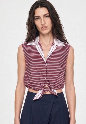 Kvinde med langt mørkt hår iført en ærmeløs bordeaux og hvidstribet bundet crop top og mørke bukser med høj talje, stående mod en hvid baggrund.
