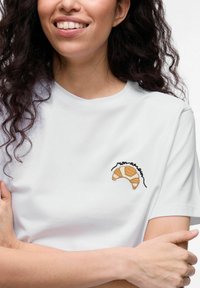 Witte katoenen t-shirt met een bruine geborduurde croissantontwerp op de linkerkant van de borst, ronde halslijn en korte mouwen.