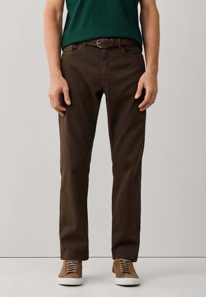 Homme portant un pantalon marron foncé, une ceinture marron, une chemise verte et des baskets en daim marron clair avec semelles blanches, debout devant un fond uni.