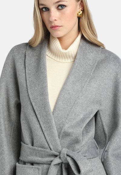 Cappotto avvolgente grigio realizzato in un tessuto morbido, con cintura da annodare, apertura frontale e maniche ampie. Indossato sopra un dolcevita color crema a coste.