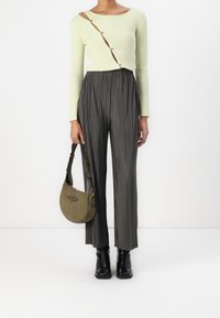 Donna che indossa un top a maniche lunghe a costine di un verde pallido con bottoni diagonali, pantaloni ampi piegati scuri, stivali neri e tiene in mano una borsa verde oliva.